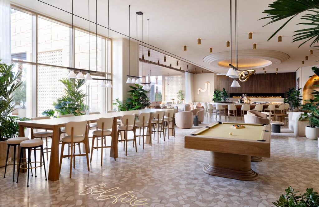 A modern, airy lounge featuring Recent Work - 11 Ravens, with large windows, neutral decor, a pool table, bar area, and cream-colored chairs. Green plants and pendant lights enhance the fresh, inviting ambiance of this stylish setting.