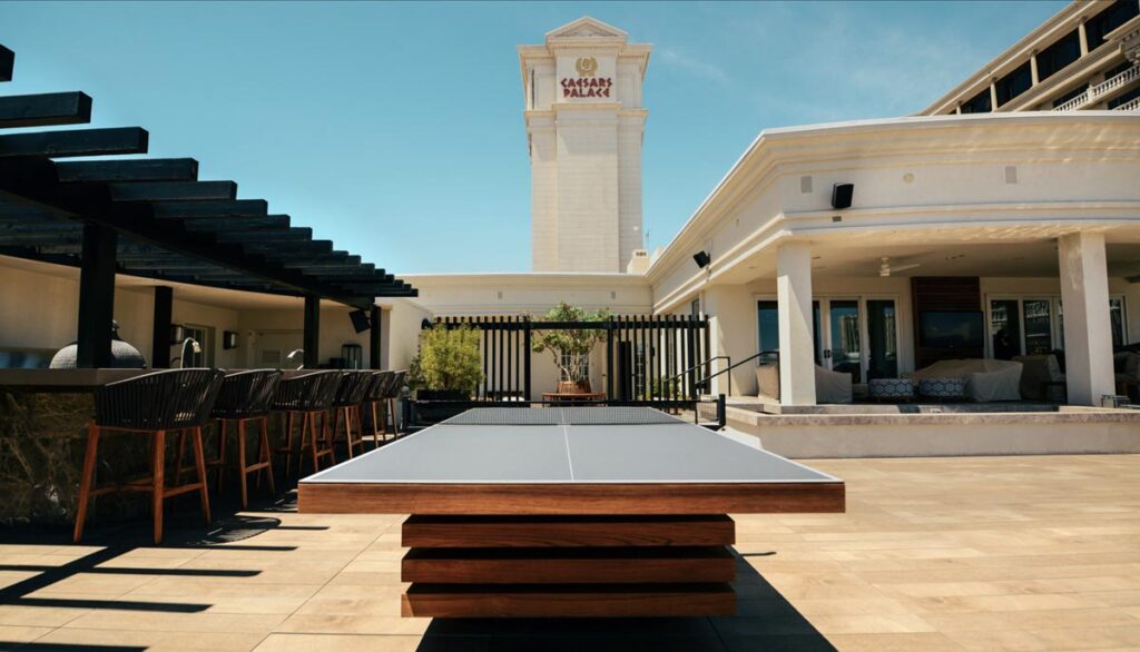 A modern outdoor patio featuring a ping pong table by 11 Ravens at its center, bar seating on the left, and a lounge area on the right. The Caesars Palace hotel tower rises in the background under a clear blue sky&mdash;showcasing recent work.