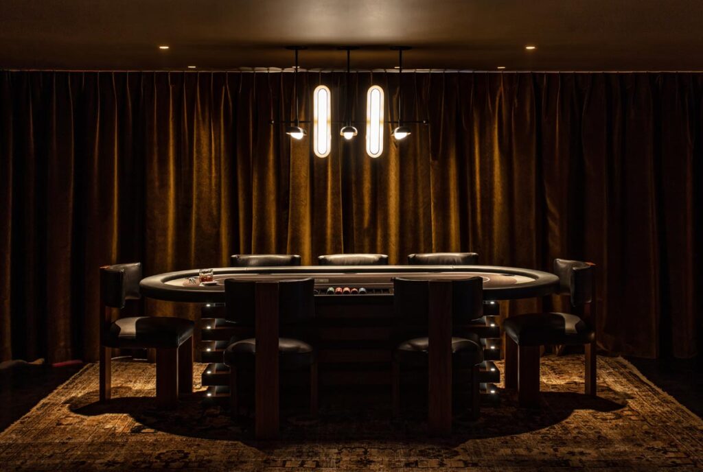 A dimly lit poker table by 11 Ravens with black chairs is centered under modern pendant lights, set against floor-to-ceiling brown curtains on a patterned rug&mdash;Recent Work that creates a moody, elegant atmosphere.