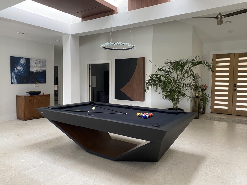 A modern room featuring a geometric black pool table by Recent Work - 11 Ravens, pool balls and cues, abstract art on white walls, a potted palm, and wooden double doors with glass panels. Natural light streams in from skylights above.