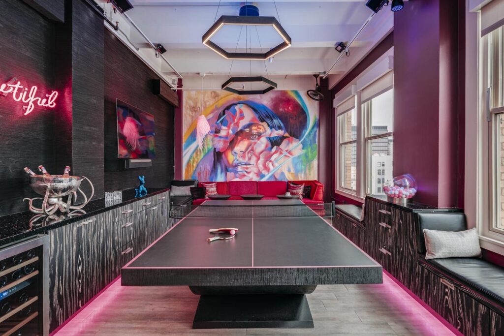Recent Work - 11 Ravens: Modern game room with a black ping pong table, vibrant wall mural of abstract faces, neon pink lighting, and contemporary decor, including a neon sign, metallic champagne bucket, and large windows with city views.