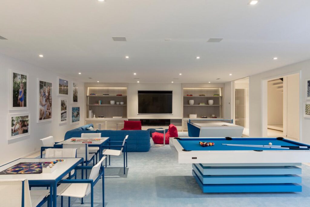 Bright modern game room featuring a blue pool table by Recent Work - 11 Ravens, game tables, blue sofa with red pillows, wall-mounted TV, open shelving, and framed photos on white walls. The space is well-lit and organized.