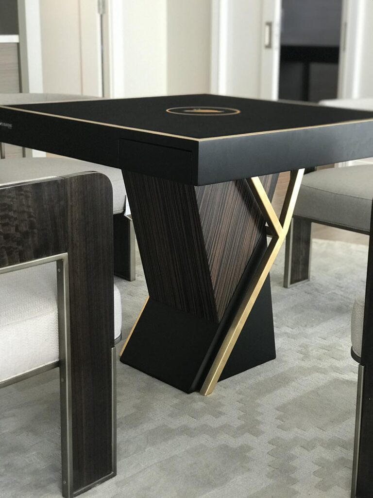 A modern square table with a black and wood finish, inspired by recent work from 11 Ravens, featuring an artistic base with intersecting gold and wood elements, surrounded by cushioned chairs on a patterned light carpet.