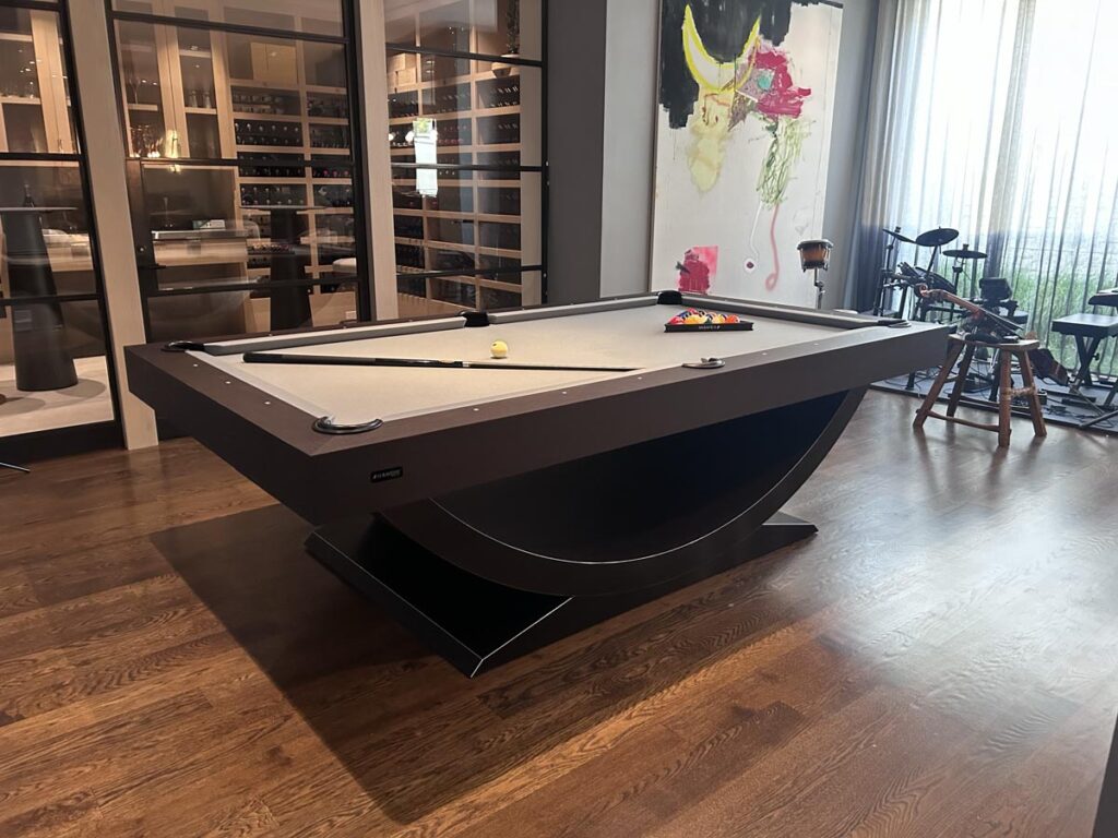 A modern pool table by 11 Ravens with a gray felt surface stands in a stylish room with wood flooring, floor-to-ceiling windows, artwork on the wall, and an electronic drum kit&mdash;showcasing recent work blending luxury and creativity.