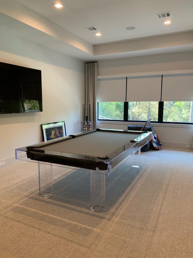 A modern, clear acrylic pool table from Recent Work - 11 Ravens sits in a carpeted room with large windows, a wall-mounted TV, pool cues in a rack, framed artwork on the floor, and a toy car by the window.