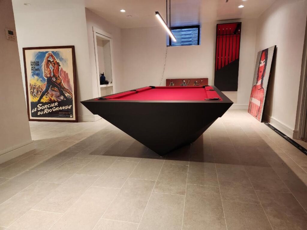 A modern room showcases a red, pyramid-shaped pool table by 11 Ravens in the center, surrounded by movie posters on the walls and floor. The space is well-lit and features light-colored tile flooring&mdash;a stunning example of recent work.
