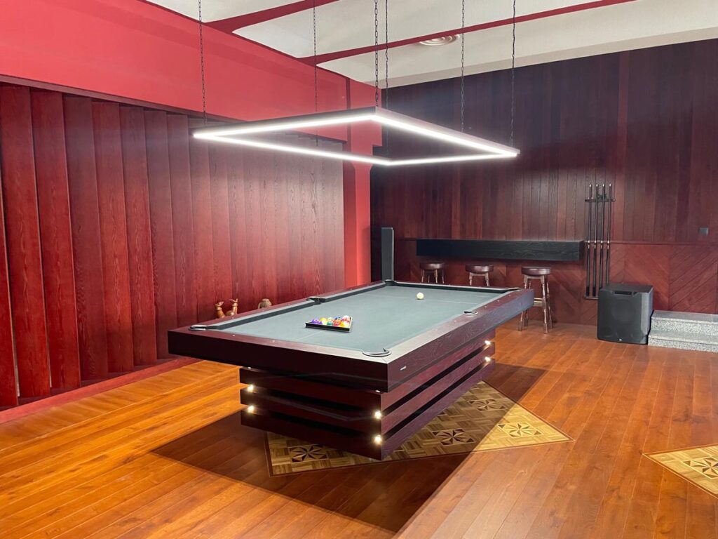 A modern billiards room by 11 Ravens features a dark wood pool table, balls set up for play, geometric overhead light, wooden floors, and red and dark panelled walls. A cue rack and stools complete this stunning example of Recent Work.