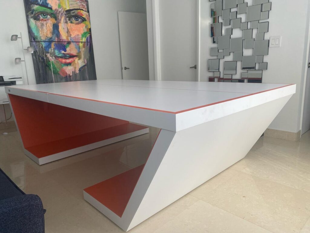 A modern, white and orange table from Recent Work - 11 Ravens with angular, geometric legs sits on a light tile floor. In the background, there is a colorful abstract portrait and a wall of square mirrors.