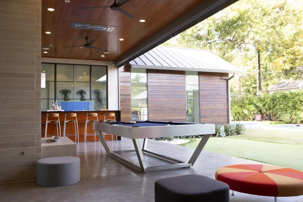 Modern indoor-outdoor recreation room featuring a sleek 11 Ravens pool table, stylish stools at a wooden bar, pouf seating, and large open windows that overlook a sunlit yard with greenery and a modern building&mdash;showcasing recent work.
