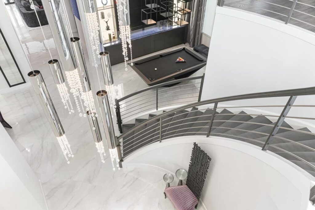 A modern, open interior features a curved staircase with metal railings, marble floors, a hanging chandelier with tubular lights, and a Recent Work - 11 Ravens pool table set up near a glass display case.