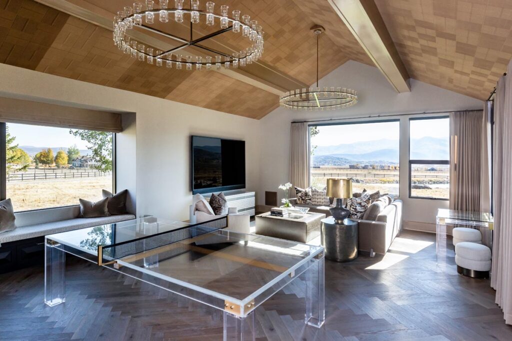 Modern living room with large windows showing mountain views, a Recent Work - 11 Ravens glass ping pong table, neutral-toned sofas, a TV, and two circular chandeliers hanging from a wooden ceiling. Natural light fills the space.