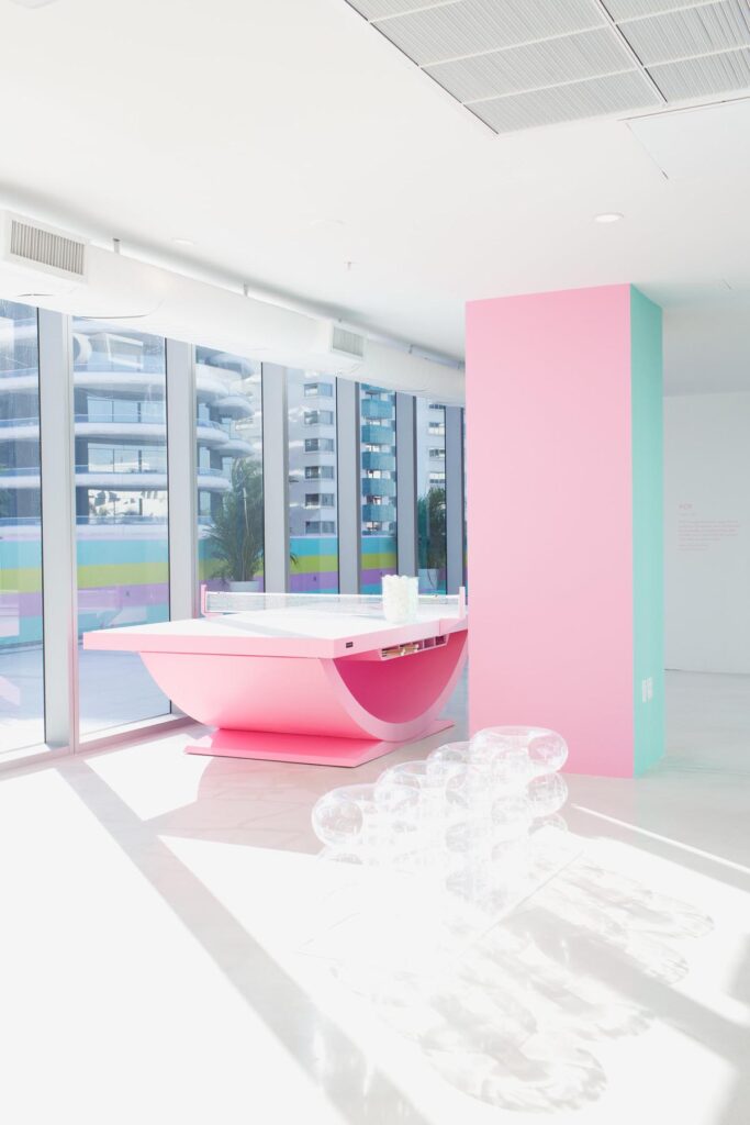 A brightly lit modern room with floor-to-ceiling windows, a large pink geometric counter, pastel blue and pink walls, and clear bubble-like chairs. Recent Work - 11 Ravens is visible through the buildings outside the windows.