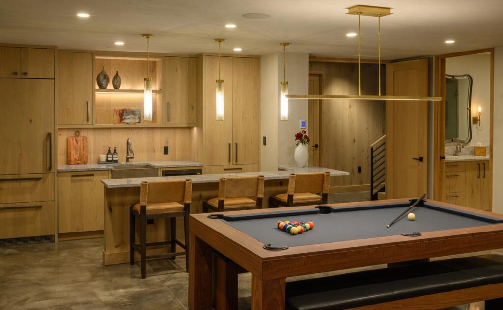 Recent Work - 11 Ravens: Modern home recreation room featuring a wooden pool table, bar stools at the kitchen island, light wood cabinetry, pendant lights, and a view into a bathroom. Pool balls and cue are perfectly arranged on the table.