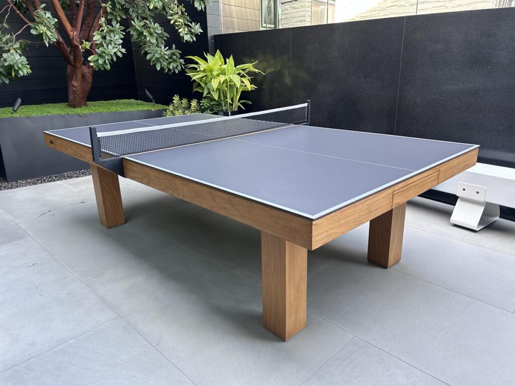 A modern outdoor ping pong table by 11 Ravens with a wooden frame and a dark playing surface, set on a light gray tiled patio with plants and a tree in the background&mdash;part of our recent work.