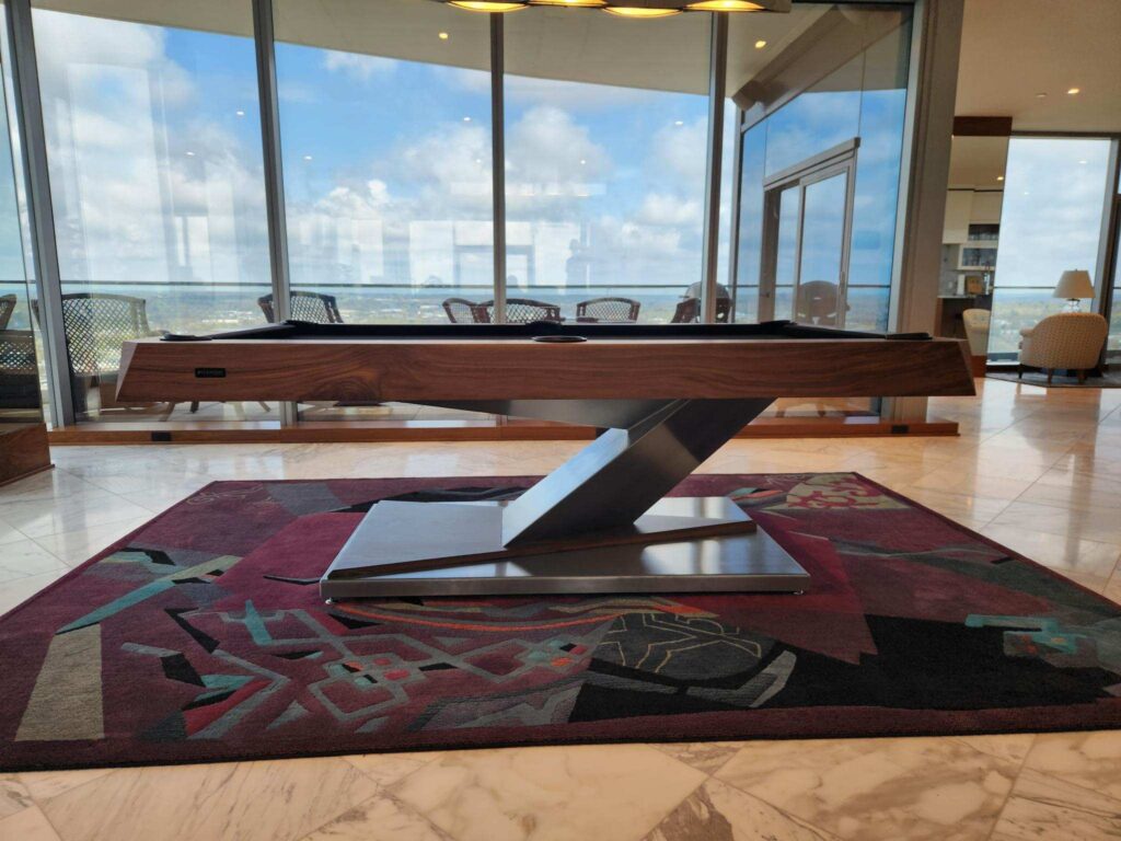 A modern pool table by 11 Ravens with a sleek wooden design sits on a patterned rug in a bright room with marble floors and large windows, showcasing this recent work against a scenic cityscape under a partly cloudy sky.