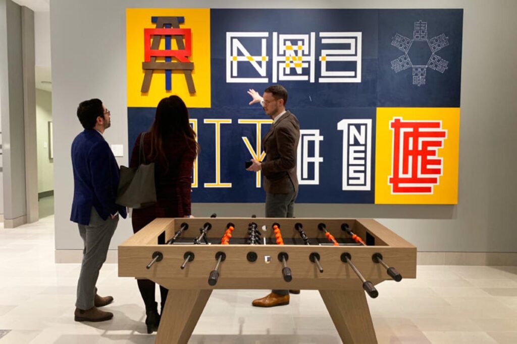 Three people stand before a colorful, geometric art piece on the wall&mdash;recent work by 11 Ravens. One gestures toward the artwork as a foosball table sits in the foreground of the modern gallery space.