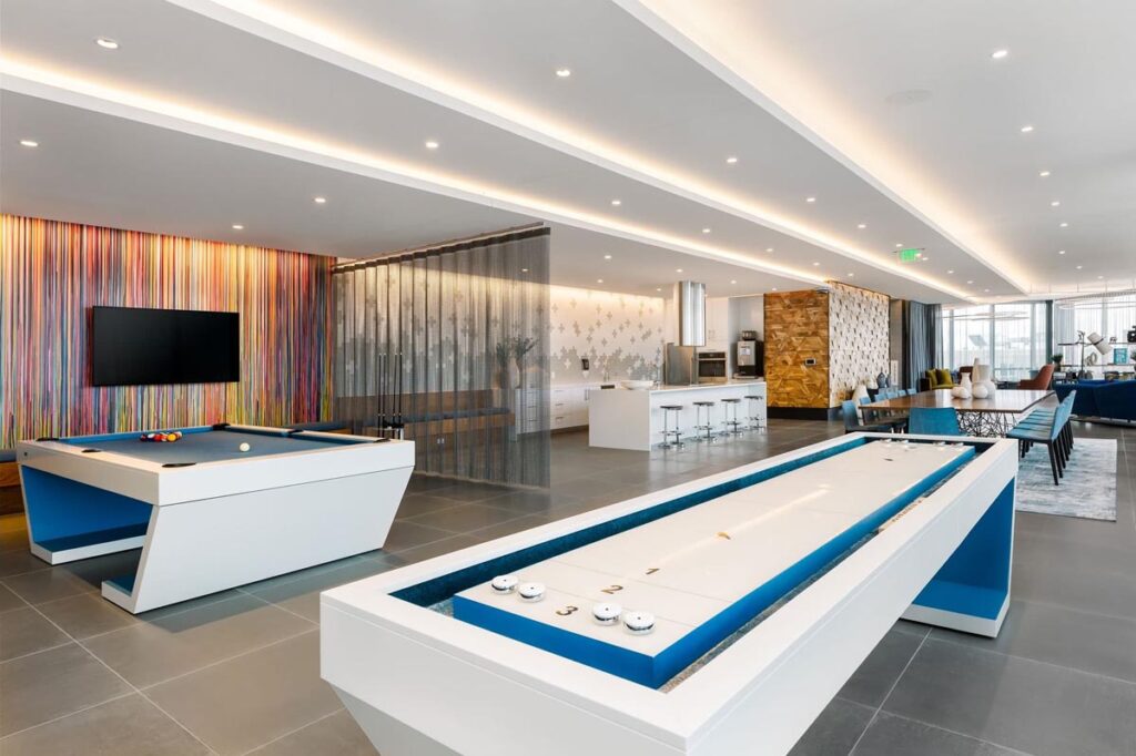 Modern game room featuring a Recent Work - 11 Ravens pool table and shuffleboard, colorful striped wall art, wall-mounted TV, bar seating, and a stylish open seating area&mdash;all illuminated by bright recessed lighting.