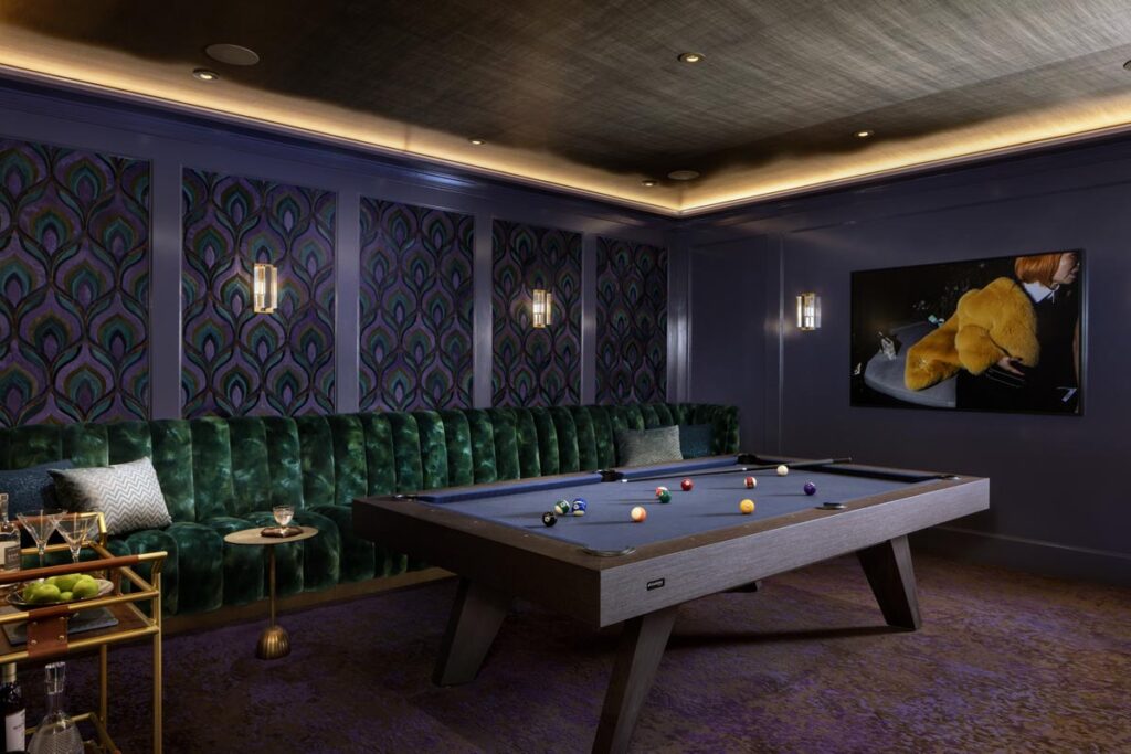 A stylish game room with a modern 11 Ravens pool table, green velvet tufted seating, patterned wall coverings, dim lighting, a wall-mounted TV, and a small side cart with drinks and fruit&mdash;one of our recent work highlights.