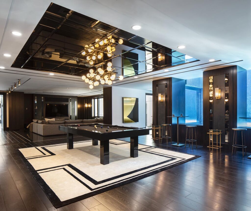 A modern game room featuring a pool table by 11 Ravens on a geometric rug, dark wooden floors, stylish lighting fixtures, a mirrored ceiling, bar area with stools, and a gray sectional sofa in the background&mdash;a showcase of recent work.