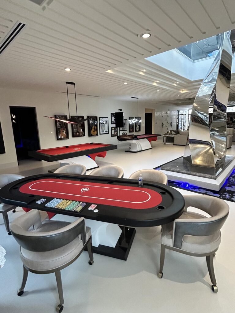 A modern game room featuring a red poker table, six chairs, poker chips, water bottle, two pool tables by Recent Work - 11 Ravens, framed guitars on the wall, and a striking mirrored sculpture over a blue-lit water feature.