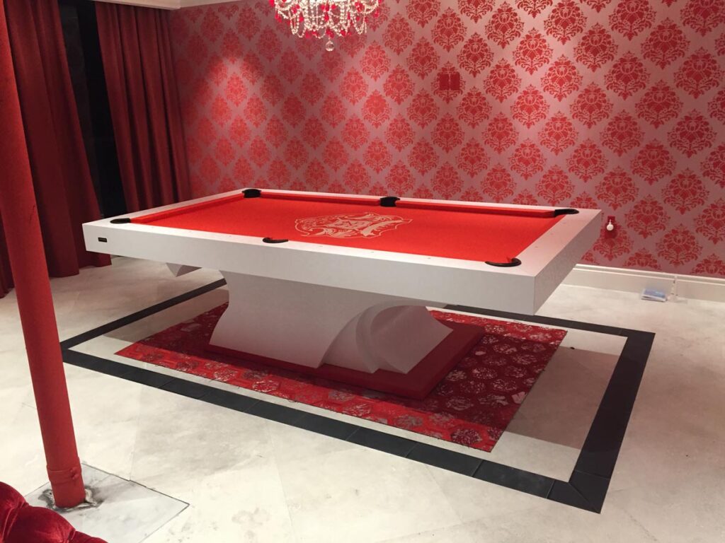 A modern pool table by 11 Ravens with a red felt surface and white frame sits on a red patterned rug in a room with red damask wallpaper, red curtains, and a red chandelier&mdash;showcasing recent work in luxury design.