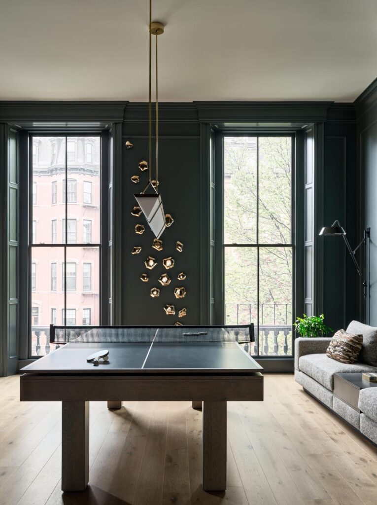 A modern living room with tall windows, a gray sofa, a floor lamp, wall art with gold elements, and a Recent Work - 11 Ravens table tennis table as the centerpiece. Light wood floors and dark green walls complete the space.