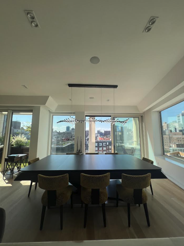 A modern dining room featuring a dark rectangular table by Recent Work - 11 Ravens, six patterned chairs, a unique hanging light fixture, large windows, and a cityscape view. Natural light fills the inviting space.