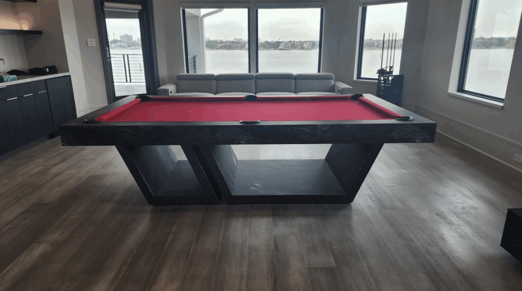 A modern pool table by 11 Ravens with a red felt top and geometric black base stands in a bright room with large windows, a gray sectional sofa, and a view of water outside&mdash;showcasing recent work at its finest.