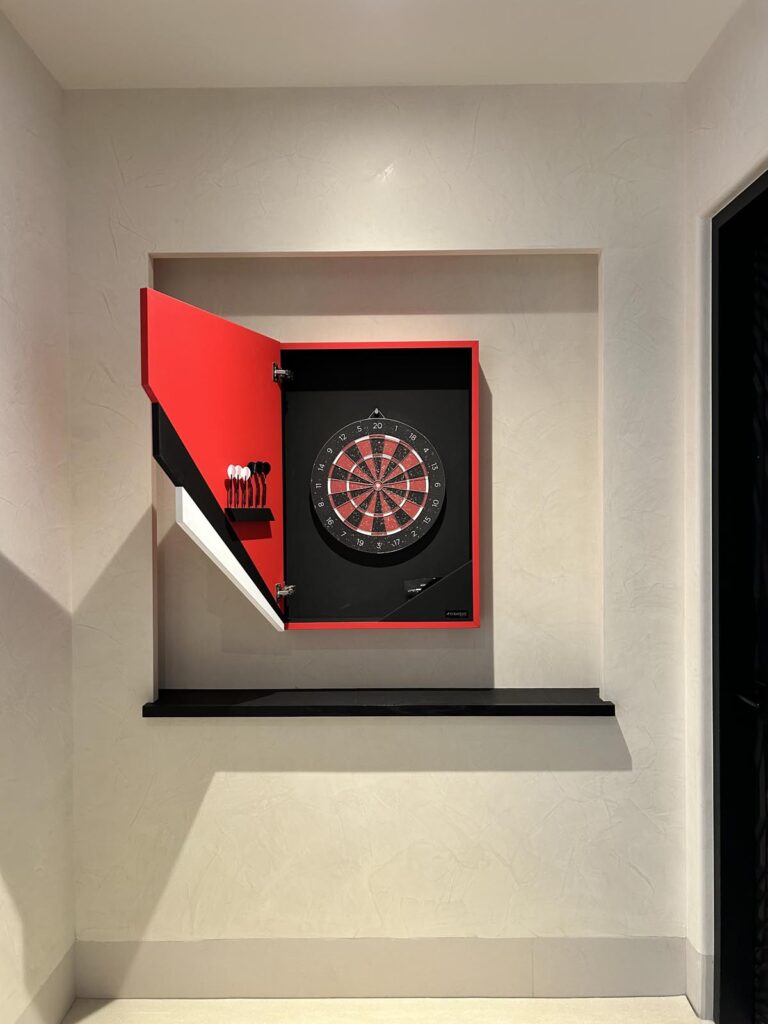 A red dartboard cabinet, part of a Recent Work - 11 Ravens collection, sits on a recessed wall shelf with its door open, showcasing a dartboard and sleek black darts inside in a brightly lit room.