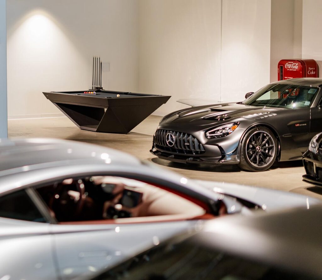 A modern showroom features a sleek black Mercedes sports car, a metallic silver car in the foreground, a geometric black pool table&mdash;Recent Work by 11 Ravens&mdash;and a red vintage Coca-Cola vending machine against a white wall.