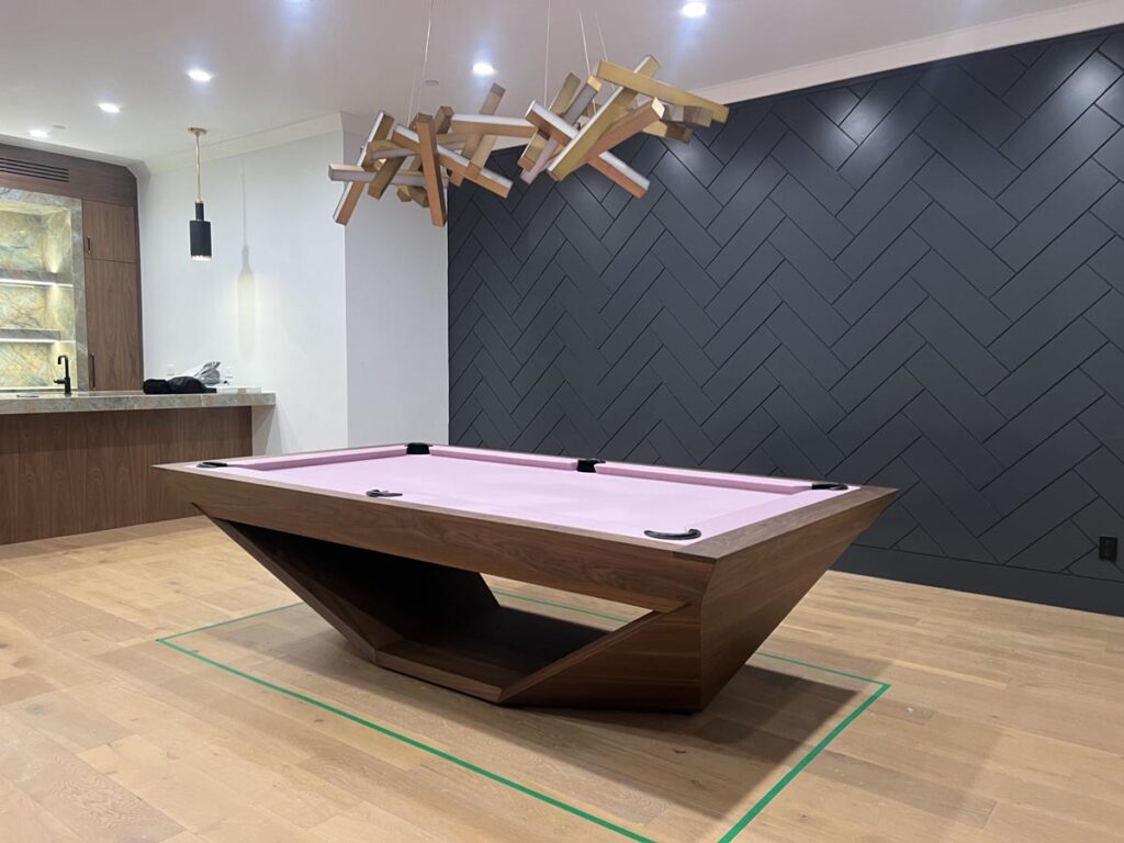 A modern room showcasing a Recent Work - 11 Ravens geometric wooden pool table with a light pink felt top, wooden flooring, a zigzag-patterned dark accent wall, unique pendant lights, and a small kitchen area in the background.