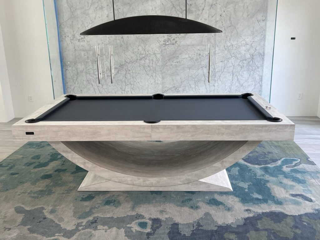 A modern pool table by 11 Ravens with a unique curved base, matte gray finish, and dark playing surface sits on a patterned blue and green rug under a sleek black light. Marble-textured wall in the background showcases this recent work.