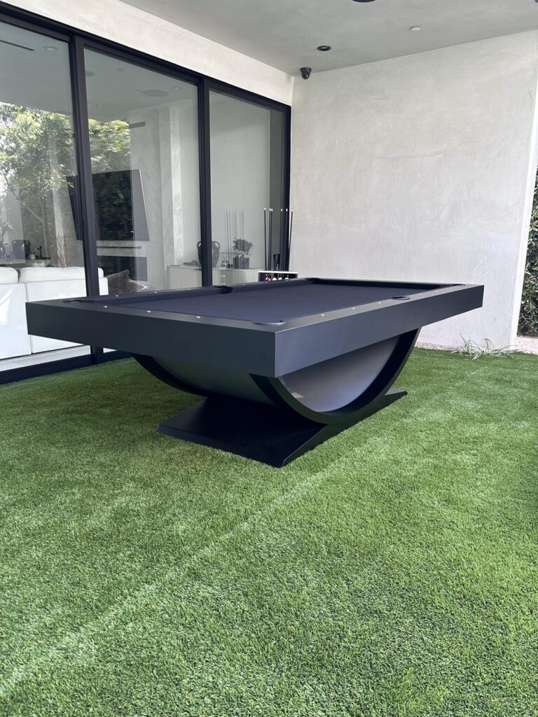 A modern black billiards table by 11 Ravens with a unique curved base stands on artificial grass in an outdoor area next to glass doors and a white wall. Pool cues and balls are visible in the background of this recent work.