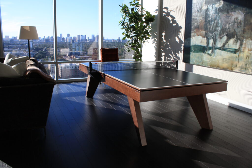 A ping pong table by Recent Work - 11 Ravens is placed in a modern living room with dark wood floors, large windows overlooking a city skyline, a floor lamp, a sofa, a plant, and a large abstract painting on the wall.