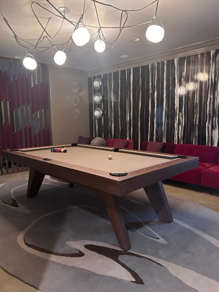 A modern room featuring a Recent Work - 11 Ravens pool table on a patterned rug, overhead lighting, a red cushioned bench along the wall, and abstract black-and-white wall art in the background. Pool balls and cue stick are arranged on the table.