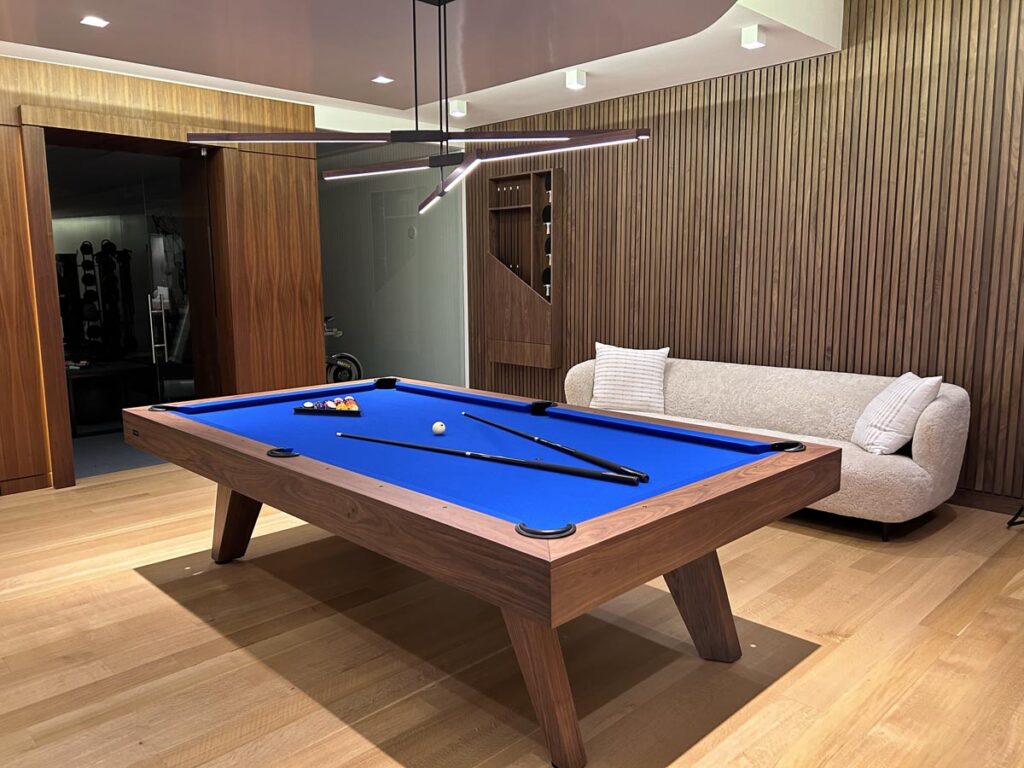 A modern room featuring a blue felt pool table by 11 Ravens, two cue sticks, and racked balls. There&rsquo;s a beige sofa with two cushions, wood panel walls, and a sleek ceiling light&mdash;showcasing recent work in luxury game room design.