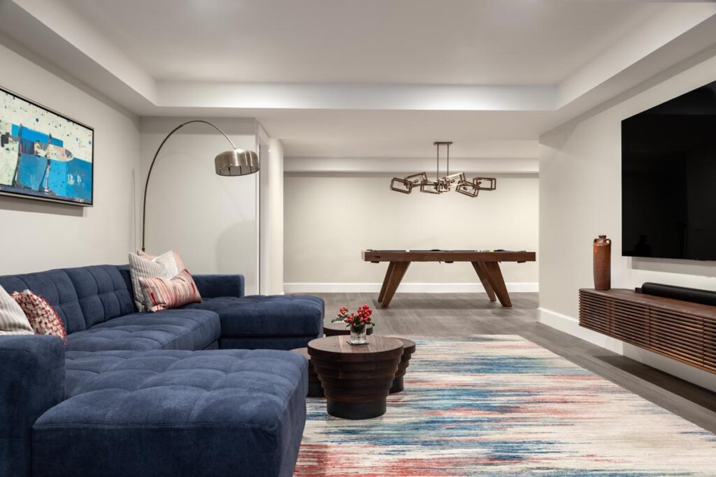 Modern living room with a navy blue sectional sofa, colorful rug, round coffee table, and large wall-mounted TV. A Recent Work - 11 Ravens pool table and unique chandelier are in the background. Artwork and decorative items complete the space.