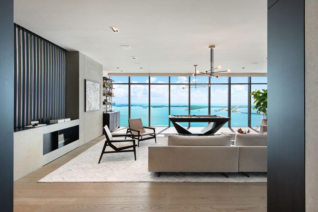 A modern living room with large floor-to-ceiling windows overlooking the ocean, featuring neutral-toned furniture, a Recent Work - 11 Ravens pool table, minimalist decor, and natural light filling the space.