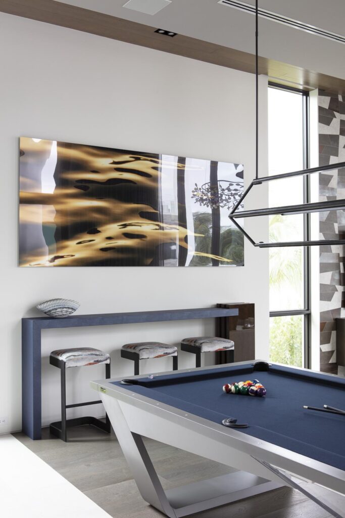 A modern room featuring a sleek 11 Ravens pool table, stools tucked under a narrow console, abstract wall art, and large windows bathed in natural light. A geometric light fixture glows overhead&mdash;an inspiring example of recent work.