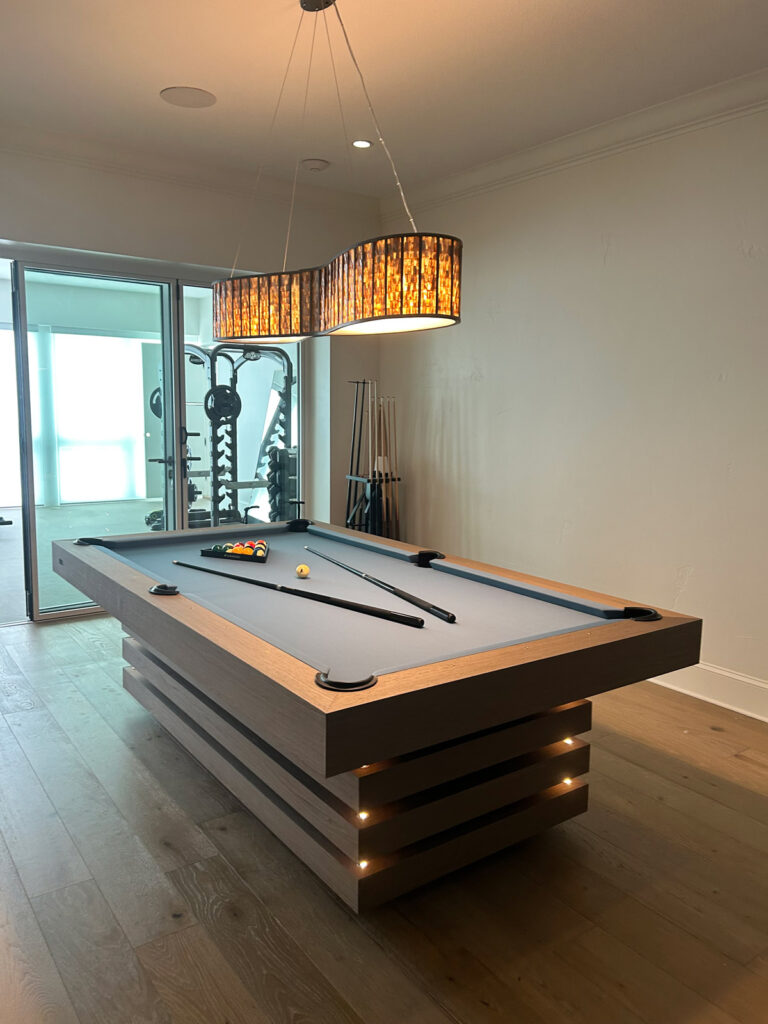 A modern game room features a light wood pool table with a blue felt top by 11 Ravens, billiard balls ready for play, two cues on the table, and a decorative pendant light above. Pool cues and gym equipment are in the background&mdash;a showcase of recent work.