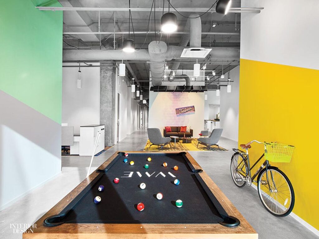 Modern office space featuring a Recent Work - 11 Ravens pool table in the foreground, a yellow bicycle to the right, colorful geometric wall accents, and a stylish lounge area with sofas and a rug in the background.