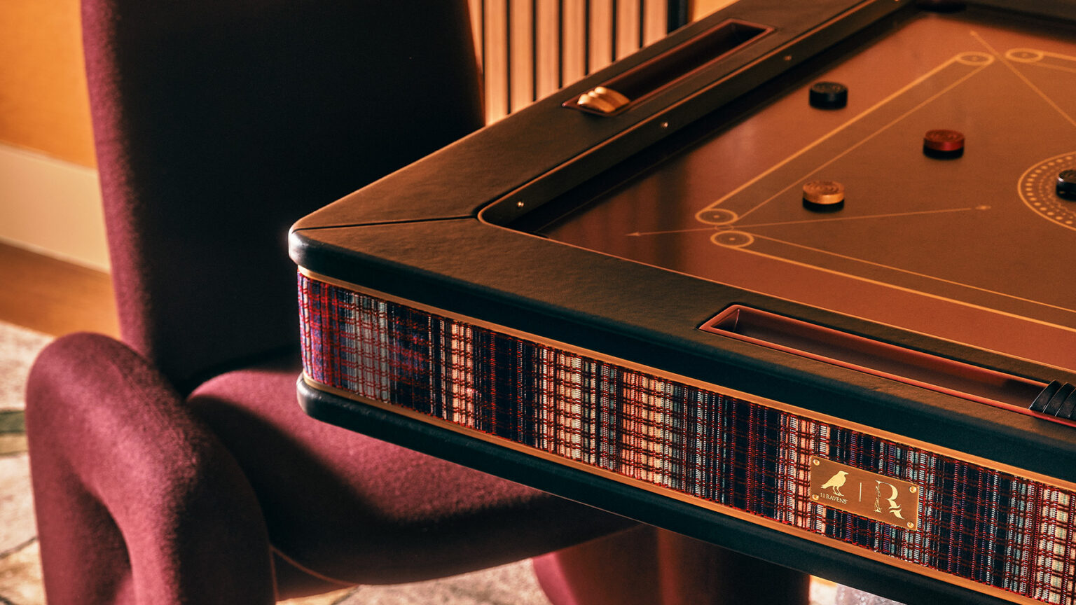 A close-up of a luxury carrom board with a patterned side panel and gold accents, placed next to a plush, burgundy chair in a warmly lit room. Handcrafted game tables like this feature arranged carrom pieces on the board’s surface.