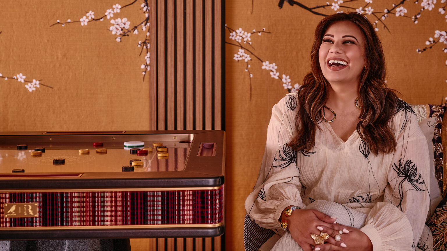 A woman with long brown hair, wearing a cream-colored blouse with black leaf patterns, sits smiling and laughing beside a luxury handcrafted game table. The background features a gold wall adorned with white cherry blossom designs.