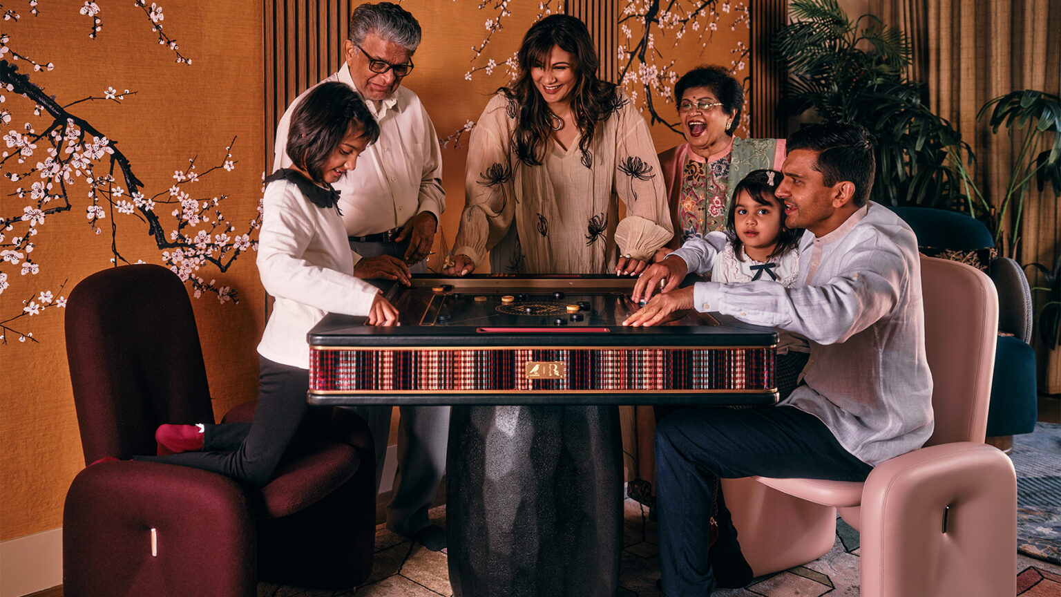 A family of six, including two young girls, smiles and gathers around a luxury game table in a warmly decorated room with floral wallpaper and modern chairs.