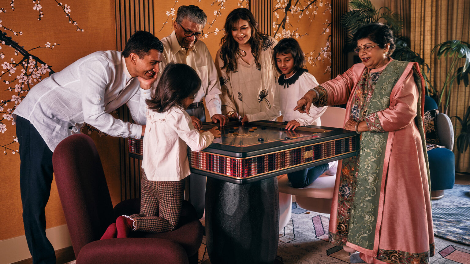 A multigenerational family gathers around a handcrafted carrom board, smiling and engaged in the game. Adults and children enjoy playing together on this luxury game table in a warmly decorated room with floral wall art.