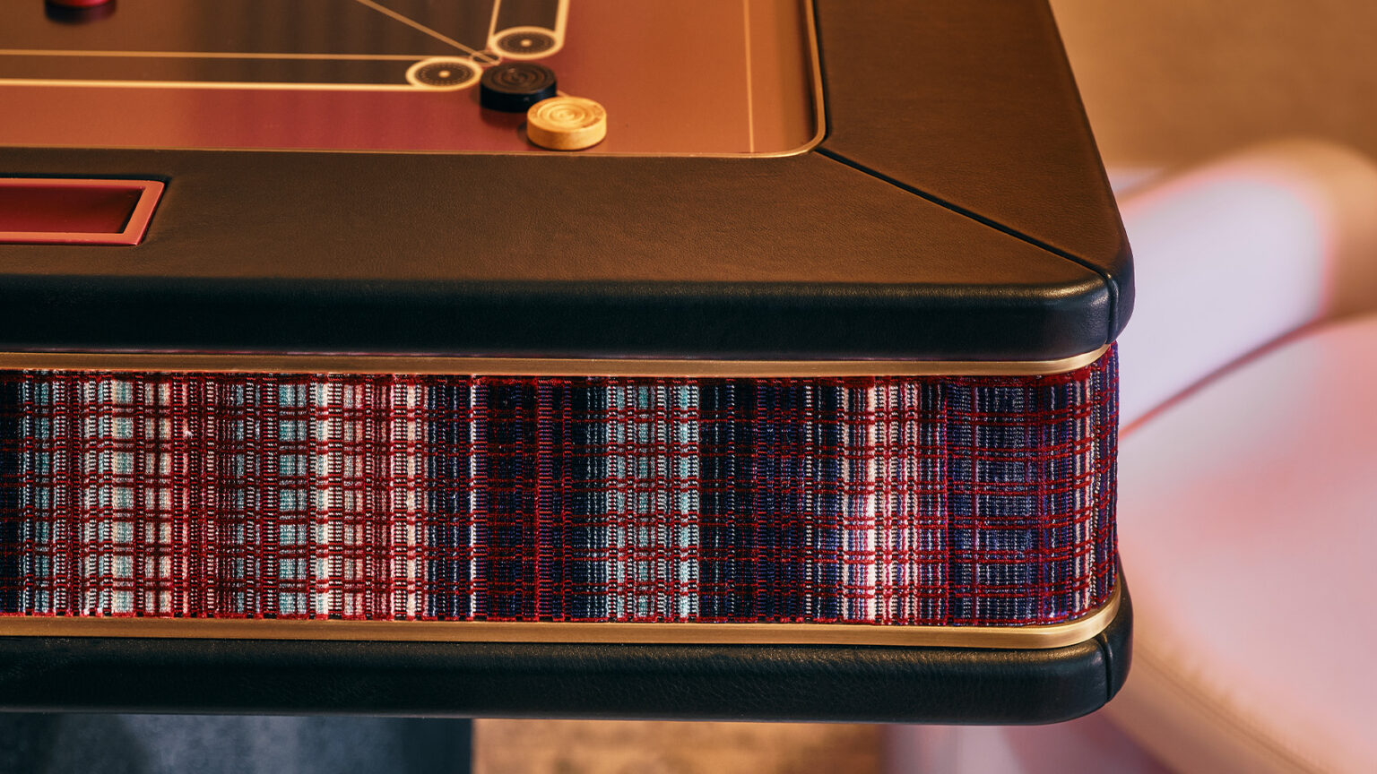 Close-up of a luxury game table with a black edge, plaid-patterned fabric side in red, blue, and white, and game pieces on top. A light-colored cushioned chair is partially visible in the background.