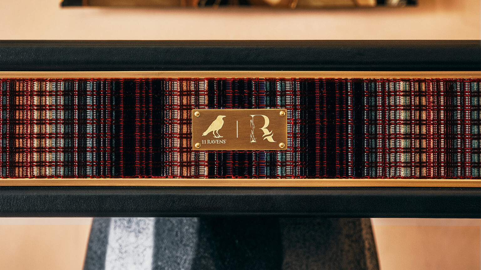 A close-up of a patterned, multicolored textile with a brass plaque in the center featuring a raven silhouette and the letter R. The luxury plaque is attached to a handcrafted wooden frame.