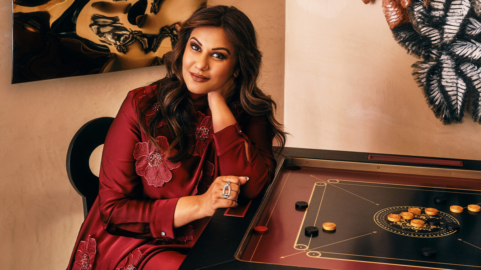 A woman in a maroon dress with floral designs sits at a handcrafted game table, resting her chin on her hand and smiling softly. The warmly lit setting features artwork on the beige walls behind her.