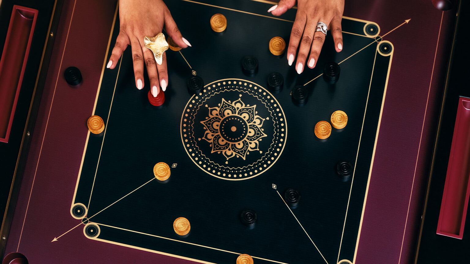 A person with manicured nails and rings moves pieces on a handcrafted black and gold carrom board decorated with a mandala design, with black and yellow tokens arranged on this luxury game table.
